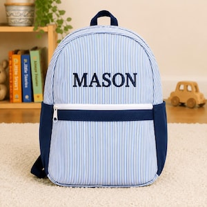 May include: A blue and white pinstriped backpack featuring a top handle, a front zippered pocket, and dark blue side panels. The name MASON is embroidered in dark blue capital letters across the center of the front panel of the backpack.