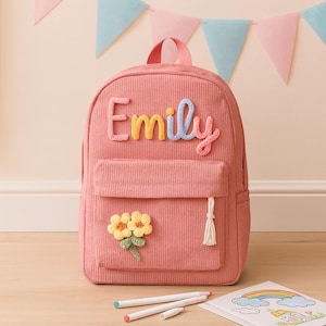 May include: A pink corduroy backpack featuring the name Emily in multi-coloured, textured yarn letters across the top. The front pocket is decorated with a yellow crocheted flower applique and a small white tassel attached to the zipper pull.