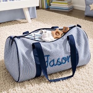 May include: A blue and white striped duffel bag with dark blue handles and the name Jason embroidered in blue script on the front. The bag is open, revealing a striped baby onesie, white socks, a wooden star toy, and a wooden block with the letter J.