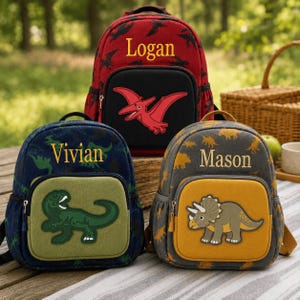 May include: Three backpacks featuring dinosaur themes and personalised names. The red bag displays 'Logan' with a pterodactyl, the blue bag shows 'Vivian' with a T-Rex, and the grey bag features 'Mason' with a triceratops. Each has a front pocket.