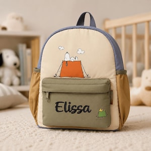 May include: A beige backpack with blue trim and tan side pockets. The front features a graphic of Snoopy on an orange tent with clouds. A green front pocket displays the embroidered name Elissa and a small graphic of Woodstock in a green tent.
