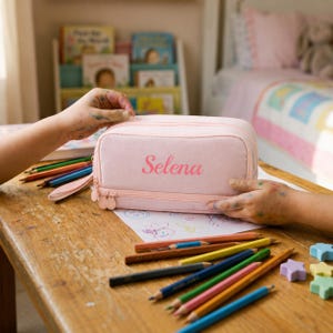 May include: A light pink rectangular pencil case with a zipper closure and the name Selena printed in a darker pink script font on the front. The case is made of fabric and features a small side handle, resting on a wooden surface with colored pencils.
