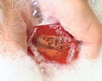 May include: A hexagonal red soap bar featuring a detailed brown embossed dinosaur skull graphic on its surface. The soap is surrounded by white bubbly foam, highlighting the contrast between the smooth red surface and the textured fossil design.