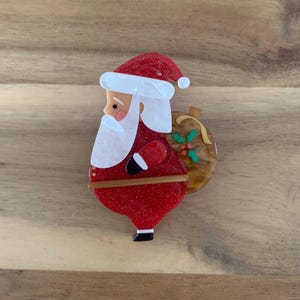 May include: A red glitter Santa Claus brooch with a white beard, hat trim, and a brown bag with holly. A festive accessory for the Christmas season.
