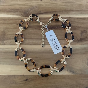 May include: A gold-toned chain necklace with oval links in a tortoiseshell pattern. The necklace has a lobster clasp closure and a tag that reads "LAUREN Ralph Lauren". The necklace is displayed on a wooden surface.