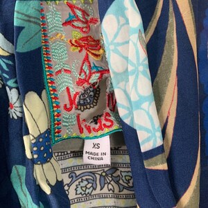 May include: Close-up of a navy blue garment with a floral pattern. The garment features an embroidered label with the text "Jus Iris" and a size tag that reads "XS". The garment is made in China.