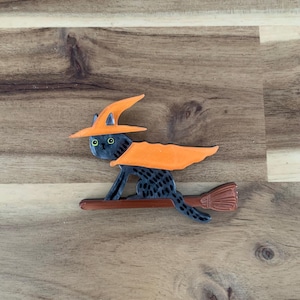 May include: A black cat wearing an orange witch hat and cape, riding a broom. The cat has green eyes and is painted with a textured pattern. The broom is brown with a reddish-brown brush. Halloween-themed decoration.