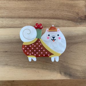 May include: A charming dog-shaped brooch in white, with a curly tail and an orange hat. The dog is adorned with a red and brown polka-dot coat, yellow trim, and carries a green gift with a red bow.