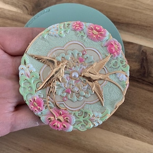 Op de afbeelding: Een ronde, decoratieve compact met een lichtgroene achtergrond en gouden accenten. De compact heeft twee gouden vogels, roze bloemen en een parel in het midden. De compact ligt op een lichtgroen oppervlak.