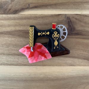 May include: A black sewing machine brooch with gold-tone decorative accents, a red top spool, and a silver-tone handwheel. A piece of pink and red patterned fabric is positioned under the needle, creating a detailed and colorful accessory.