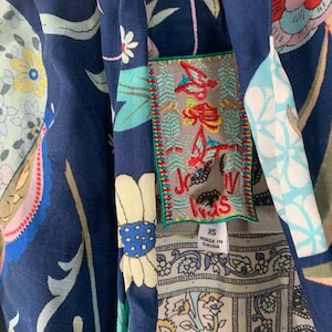 May include: Close-up of a navy blue garment with a floral pattern in shades of pink, yellow, and white. A decorative patch with embroidered text and a size tag is visible. The garment has a unique, artistic design.