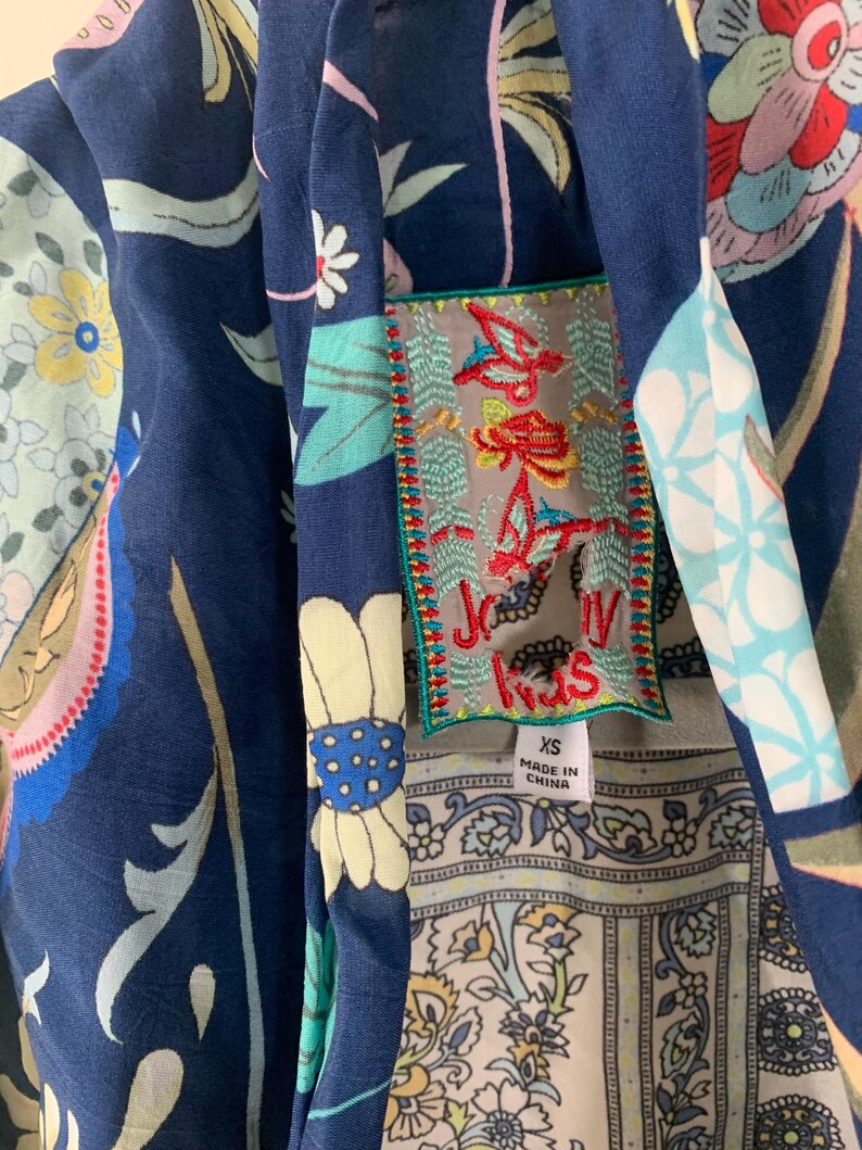 May include: Close-up of a navy blue garment with a floral pattern in shades of pink, yellow, and white. A decorative patch with embroidered text and a size tag is visible. The garment has a unique, artistic design.