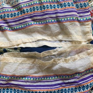 May include: Close-up of a section of a patterned fabric. The fabric features horizontal stripes in purple, teal, and orange against a white background. The bottom edge has a cream-colored trim. The fabric is slightly wrinkled.