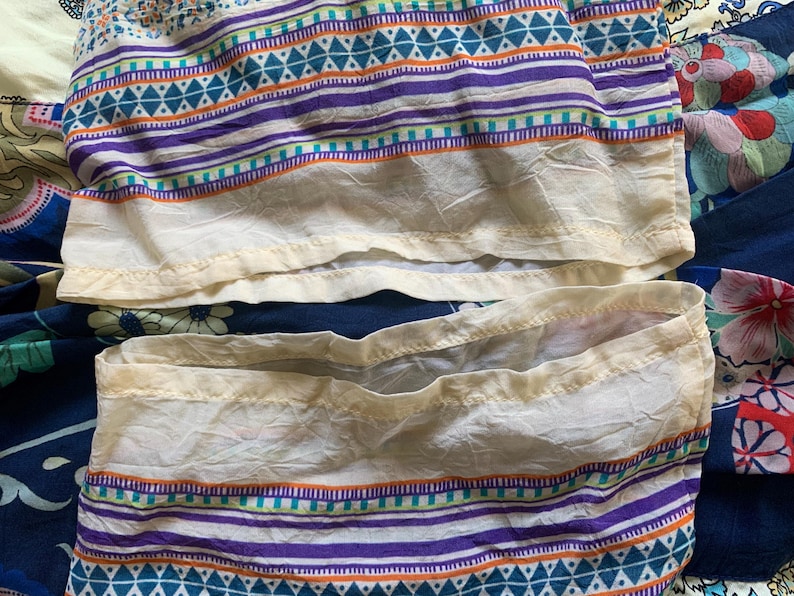 May include: Close-up of a section of a patterned fabric. The fabric features horizontal stripes in purple, teal, and orange against a white background. The bottom edge has a cream-colored trim. The fabric is slightly wrinkled.