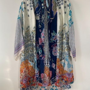 May include: A long, patterned kimono in shades of white, navy blue, orange, and purple. The kimono features a variety of floral and paisley designs. It has long sleeves and a tie closure at the neck, creating a flowing, bohemian style.