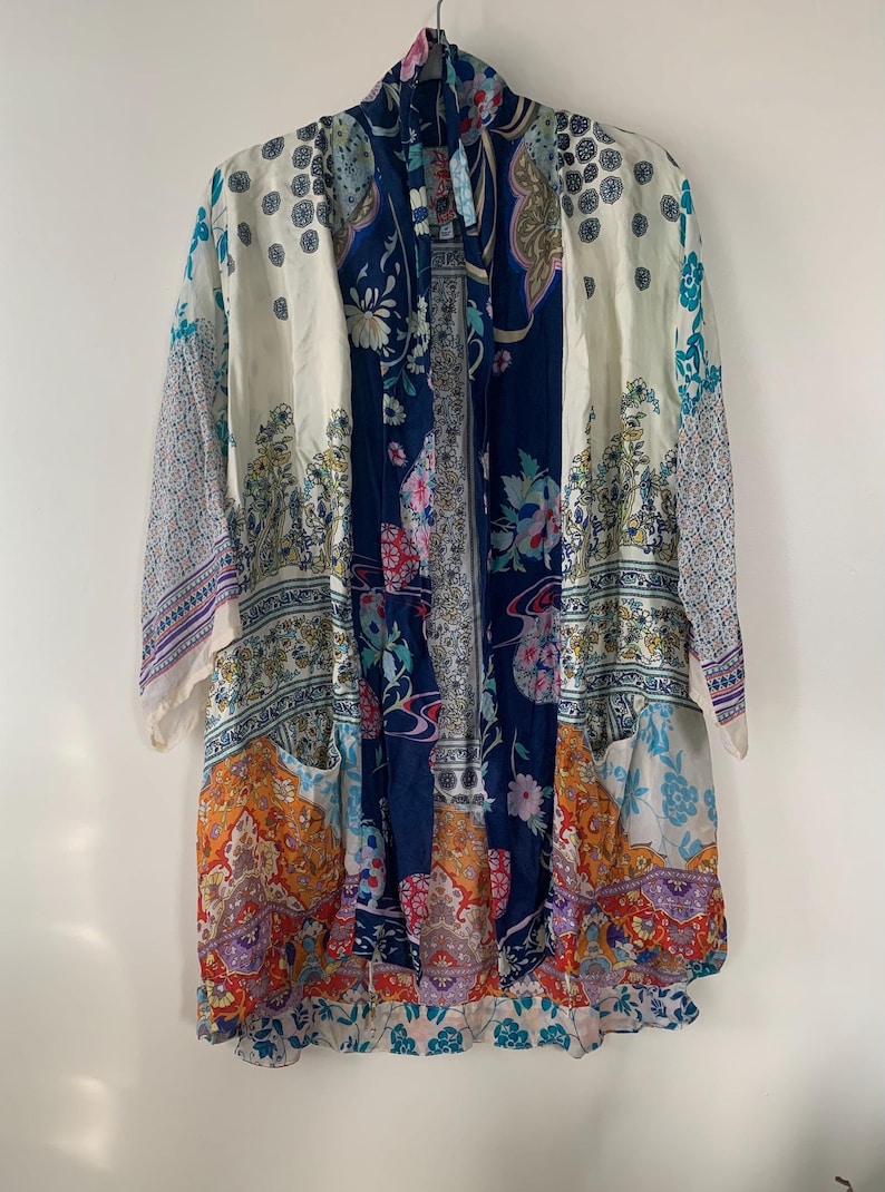 May include: A long, patterned kimono in shades of white, navy blue, orange, and purple. The kimono features a variety of floral and paisley designs. It has long sleeves and a tie closure at the neck, creating a flowing, bohemian style.