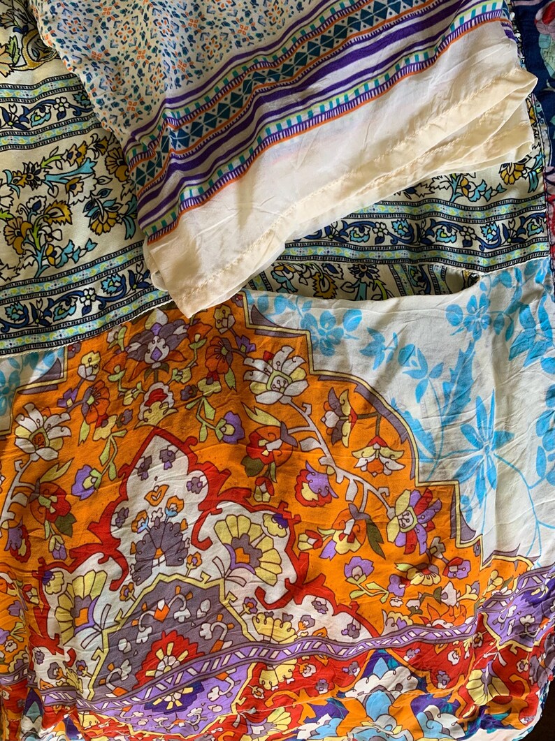 May include: A collection of colorful fabric pieces with intricate floral and geometric patterns. The fabrics feature a mix of orange, blue, purple, and cream colors, creating a vibrant and artistic display. The textiles appear to be lightweight.