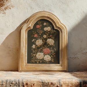 Op de afbeelding: Een ingelijste bloemenprint met witte en roze rozen op een donkere achtergrond. De lijst is van licht, verweerd hout met een gewelfde bovenkant. Het kunstwerk wordt tentoongesteld op een getextureerd oppervlak.