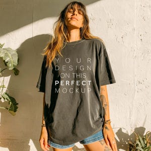 May include: A dark gray t-shirt with the text "YOUR DESIGN ON THIS PERFECT MOCKUP" in white. The shirt has a crew neck and short sleeves. The model is wearing blue jean shorts.