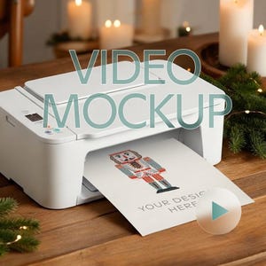 May include: A white printer is printing a card with a nutcracker design. The card has the text "YOUR DESIGN HERE". The image has a video mockup overlay. The scene is set on a wooden surface with candles and greenery in the background.
