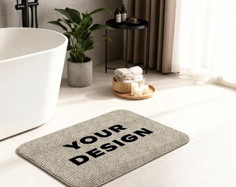 May include: A rectangular beige bath mat with the words "YOUR DESIGN" in bold black letters. The mat has a textured surface and is placed on a white tiled floor. The bathroom setting includes a white bathtub, a potted plant, and a small side table.