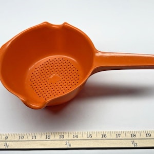 May include: An orange plastic colander with a handle. The colander has a perforated bottom and a pouring spout. The handle has a hole for hanging. The colander is approximately 10 inches long.