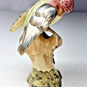 Puede incluir: Una figura de cerámica de una cacatúa galah posada sobre un tocón de árbol marrón. El ave presenta una cabeza rosada con marcas oscuras, alas de color verde claro con detalles negros y un pecho blanco. La base tiene tonos verdes y marrones.