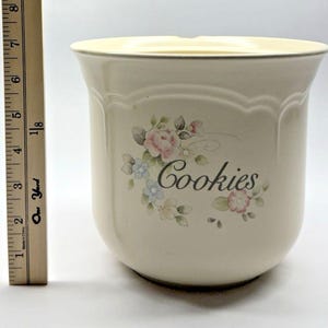 Pode incluir: Pote de biscoitos de cerâmica creme com um design floral e a palavra "Cookies". O pote tem uma borda ondulada e uma forma ligeiramente alargada. O design floral inclui flores rosa, azuis e verdes. Altura aproximada 18 cm.