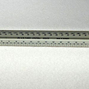 Puede incluir: Una regla de escala arquitectónica gris y blanca con medidas en centímetros. La regla está marcada con números y líneas para mediciones precisas. El texto "A.W. Faber-Castell 553/22 Made in Germany" está impreso en la regla.