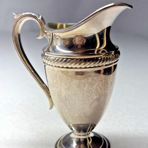 May include: A polished silver-plated creamer pitcher featuring a flared spout, a curved handle, and a pedestal base. The body is decorated with a textured, etched pattern and a prominent rope-style band encircling the upper section of the vessel.