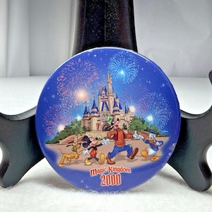 Może przedstawiać: Okrągła niebieska przypinka z grafiką zamku Kopciuszka w nocy z fajerwerkami, Plutonem, Myszką Miki, Goofym i Kaczorem Donaldem. Napis Magic Kingdom 2000 jest wydrukowany na czerwono i żółto u dołu tej okrągłej przypinki.
