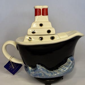 Puede incluir: Una tetera de cerámica con forma de barco de vapor con casco negro, cubierta blanca y chimenea con parte superior roja. La base presenta olas pintadas en azul y blanco, y la cubierta incluye pequeños ojos de buey circulares. Una etiqueta azul oscuro está unida al asa.