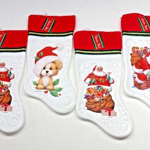 Puede incluir: Cuatro calcetines navideños blancos con puños rojos y ribete verde. Dos calcetines muestran a Santa Claus con un saco de regalos, uno muestra un cachorro con gorro de Santa y otro muestra a Santa sobre una chimenea. Tienen etiquetas Santa Best.