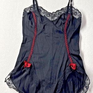 May include: A black bodysuit featuring thin spaghetti straps, a scalloped lace neckline, and two vertical red lace panels running down the front. The garment includes small red ribbon bows at the hips and black lace trim along the leg openings.