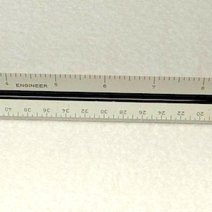 May include: A white and black architectural scale ruler with markings and text. The ruler has a black center and is marked with numbers and lines for precise measurements. The text includes "TACH", "ENGINEER", and "Germany".