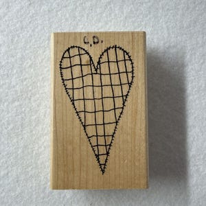 Op de afbeelding: Een rechthoekige houten rubberstempel met een zwarte inktillustratie van een hart met een ruitpatroon en gestikte randen. De letters C.D. zijn in zwarte inkt boven het hartontwerp op het lichtbruine houten blok gedrukt.