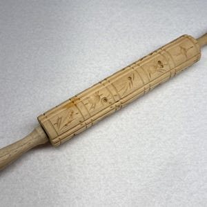 May include: A light brown wooden rolling pin featuring a repeating engraved pattern of small birds and floral elements across its cylindrical surface. The tool has two smooth, tapered handles on either end for gripping during use in the kitchen.