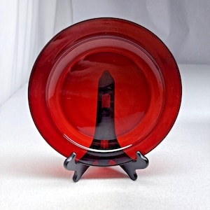 Puede incluir: Un plato circular de vidrio rojo rubí translúcido con una superficie lisa y pulida y un borde ancho y plano. El plato se mantiene en posición vertical mediante un soporte de exhibición de plástico negro de tres puntas que sostiene la base.