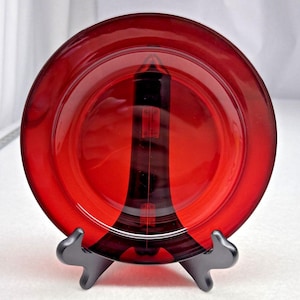 May include: A circular, translucent ruby red glass plate featuring a central embossed design of the Eiffel Tower. The plate is displayed upright on a black plastic stand, showcasing the deep red colour and the clear, moulded architectural details.