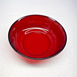 May include: A circular bowl made of translucent red glass. The bowl features a smooth, polished surface with a consistent deep crimson hue throughout the material, a wide open top, and a rounded base that tapers slightly toward the center.