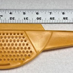 May include: A yellow plastic kitchen strainer with a handle and a perforated semi-circular bowl. A metal ruler placed above the tool indicates a total length of approximately 26.7 centimetres from the edge of the bowl to the end of the handle.