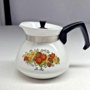 May include: A white teapot with a black handle and lid, featuring a metallic silver band around the neck. The body is decorated with a colorful botanical illustration of fruits and flowers, with the text Le Thé printed in black script below.