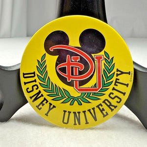 May include: A circular yellow pin featuring a black Mickey Mouse silhouette icon, a red stylized DU logo, and green laurel leaves. The text DISNEY UNIVERSITY is printed in black capital letters along the bottom edge of the circular yellow surface.