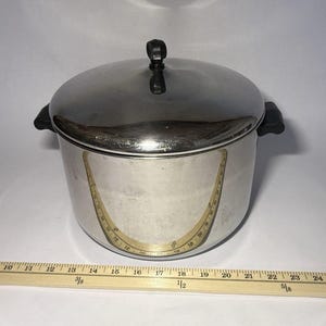 May include: A stainless steel pot with a lid and black handles. The pot has a curved measuring guide on the side. A wooden ruler is placed below the pot. The pot measures approximately 23 cm in diameter.