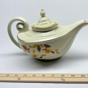 Może przedstawiać: Kremowy ceramiczny imbryk z zakrzywionym uchwytem i wylewką. Imbryk ma złote wykończenie i kwiatowy wzór w odcieniach czerwieni, żółci i brązu. Pokrywka ma ozdobne zwieńczenie. Drewniana linijka znajduje się na pierwszym planie.