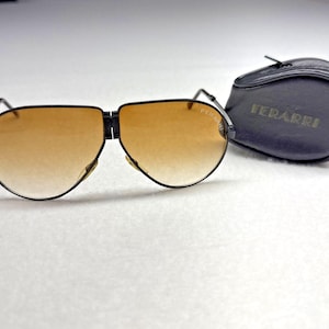 May include: Black aviator sunglasses with gradient amber lenses and the brand name FERARRI printed on the right lens. The sunglasses feature a folding bridge design and are accompanied by a black, oval-shaped protective case with the name FERARRI embossed in gold lettering on the front.