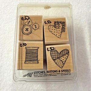 May include: A set of four wooden rubber stamps in a clear plastic case. The stamps feature black ink illustrations of buttons, a spool of thread, and two heart shapes. The text on the case reads STAMPIN UP! STITCHES, BUTTONS & SPOOLS.