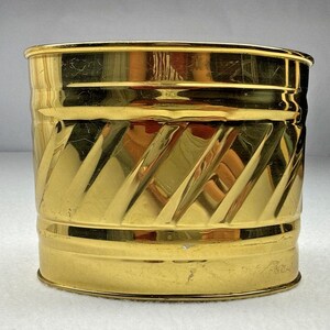Puede incluir: Una maceta de metal en tono dorado pulido con forma cilíndrica, que presenta una banda decorativa de crestas diagonales en relieve alrededor del centro y secciones horizontales lisas en los bordes superior e inferior del recipiente.