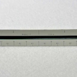 May include: A gray architectural scale ruler with black detailing and markings. The ruler is marked with measurements in inches and features the text "PARIS USA". This tool is used for architectural and engineering drawings.