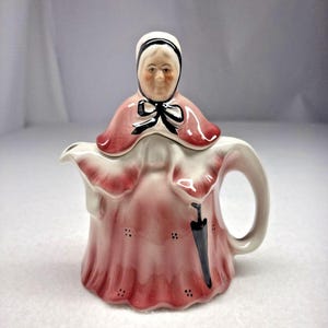 May include: A ceramic teapot shaped like a person wearing a pink dress and a white bonnet with a black bow. The figure has a painted face, a handle on the right side, and a black umbrella graphic on the front of the skirt. The teapot is glazed.